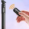 A hand holding a black Ugreen Bluetooth remote control, with a black selfie stick visible on the left. Orange signal waves emanate from the remote towards the selfie stick. Náhled obrázku produktu