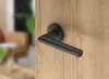 A black door handle is mounted on a wooden door. The background is blurred and shows a living room setting. Produktbild-Vorschau