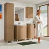 A woman in a bathrobe stands in a modern bathroom next to a sink and cabinets, looking out a window. Náhled obrázku produktu