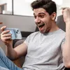 A man is sitting on a couch, holding a smartphone and looking at it with excitement. Produktbild-Vorschau