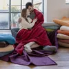 A couple is cuddling under a large red blanket, surrounded by stacks of colorful blankets. Produktbild-Vorschau
