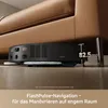 A black robot vacuum cleaner is shown cleaning under a brown leather sofa, with a measurement indicating its low profile. Produktbild-Vorschau