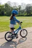 A young girl in a blue helmet stands with a blue and white bicycle in a park. Produktbild-Vorschau