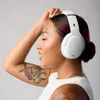 A woman wearing white headphones and a white tank top, with tattoos on her arm, poses against a white background. Náhled obrázku produktu