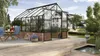 A greenhouse with plants inside, set in a backyard with a wooden deck, pond, and house in the background. Náhled obrázku produktu