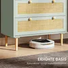 A robot vacuum cleaner is cleaning under a light green cabinet with rattan-style drawers. Produktbild-Vorschau