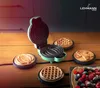 A mini waffle maker with various waffle plates and finished waffles on a wooden table. Náhled obrázku produktu