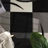 A close-up view of a rug with a geometric pattern, featuring squares and curved lines in black, gray, and white. A green leaf and a draped blanket are also visible. Anteprima dell’immagine prodotto