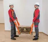 Two men in work clothes are carrying a chest using lifting straps. Produktbild-Vorschau