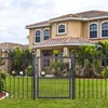 A gray metal garden gate is shown in front of a tan house with a tiled roof. The gate is partially open, revealing a well-maintained lawn and some tropical plants. The sky is overcast. Produktbild-Vorschau