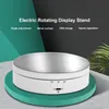 A white electric rotating display stand with a silver top surface is shown on a teal circular base against a dark background with green and black geometric shapes and a hint of greenery on the right. Náhled obrázku produktu