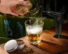 A hand is holding a wooden smoking lid over a glass of whiskey, with a smoking gun and a tin of wood chips nearby. Voir les images du produit