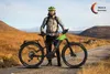 A man stands with an electric mountain bike in a natural, hilly landscape. Produktbild-Vorschau