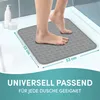 A person's feet are stepping onto a square gray shower mat in a shower stall. There are bottles of toiletries in the background. Produktbild-Vorschau