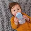A baby lying on a gray knitted blanket, drinking from a blue sippy cup with handles. Náhled obrázku produktu