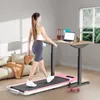 A woman is walking on a pink treadmill while working at a standing desk in a home office setting. Produktbild-Vorschau
