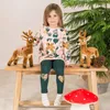 A young girl sits on a wooden bench with two stuffed deer toys, in front of a Christmas tree. Anteprima dell’immagine prodotto