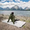 A person sits on a blanket by a lake with mountains in the background. Produktbild-Vorschau