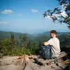 A young man sits on a wooden bench on a mountain overlook, with a backpack next to him, gazing at the scenic view. Anteprima dell’immagine prodotto