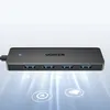 A black Ugreen USB hub with four USB ports is shown against a light gray and white background with glowing lines and icons emanating from it. Produktbild-Vorschau