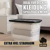 A clear plastic storage box filled with folded blankets and towels, placed on a wooden floor next to a nightstand and bed in a bedroom setting. Produktbild-Vorschau