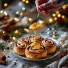 A hand is drizzling a yellow sauce over cinnamon rolls on a plate, with festive lights in the background. Produktbild-Vorschau