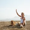 A woman and her dog are sitting on a hill with mountains in the background. Produktbild-Vorschau