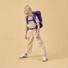 A young girl wearing a backpack and overalls, posing against a plain background. Produktbild-Vorschau