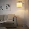 A floor lamp illuminates a living room scene with a sofa and artwork. Produktbild-Vorschau