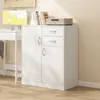 A white cabinet with two doors and three drawers, placed next to a desk with a computer monitor and books. Náhled obrázku produktu