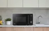 A black microwave is displayed on a kitchen counter, with cabinets above and wooden cabinets below. A sink and faucet are to the right, and a small plant and fruit basket are to the left. Produktbild-Vorschau
