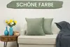 A living room scene with a sofa, pillows, a side table with vases and books, and a blanket. Produktbild-Vorschau