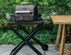 A grill stand with a grill on it, next to a wooden table, with greenery in the background. Náhľad obrázku produktu