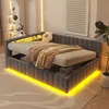 A modern bedroom with a dark gray upholstered bed featuring built-in LED lighting and storage. The bed is made up with beige bedding and a brown throw blanket. A small, white, flower-shaped pillow is on the bed. Náhled obrázku produktu