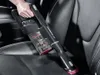 A person is using a black and red handheld vacuum cleaner to clean the dark leather seat of a car. Voir les images du produit
