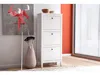 A white shoe cabinet with three drawers stands next to a floor lamp and an umbrella stand in a room with an orange wall and a window with blinds. Produktbild-Vorschau