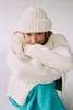 A young woman wearing a white knitted sweater and hat, with her arms crossed, looking at the camera. Produktbild-Vorschau