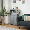 A gray cabinet is shown in a living room setting next to a sofa and a plant. Náhled obrázku produktu