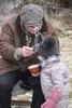 An older man is feeding a young child from a red bowl outdoors in a winter setting. Produktbild-Vorschau