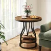 A round wooden side table with a metal frame, set next to a green armchair, with a vase of flowers, a cup of coffee, and some pastries on top. Books are stored on the lower shelf. Produktbild-Vorschau