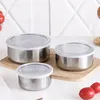 Three stainless steel food containers with clear lids are arranged on a wooden board and a white surface, with some tomatoes and nuts in the background. Produktbild-Vorschau