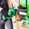 A person wearing black gloves is using a green mini chainsaw to cut a piece of wood held in a vise. Produktbild-Vorschau