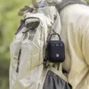 A black portable speaker with a carabiner hook is attached to a tan backpack. The background is blurred green foliage. Voir les images du produit