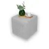 A light gray cube-shaped ottoman sits on a white background. A wooden tray with a green vase of eucalyptus leaves and a white teacup and saucer is placed on top of the ottoman. Produktbild-Vorschau