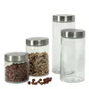 Four glass storage jars with silver lids are arranged on a white background. Two jars contain food items (nuts and granola), and two are empty. Some nuts are scattered in front of the jars. Náhled obrázku produktu