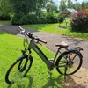 An electric bicycle is parked on a grassy area next to a paved path in a park. Produktbild-Vorschau