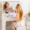 A woman is sitting at a white vanity table and drying her hair in front of a mirror. Produktbild-Vorschau