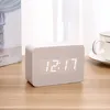 A digital alarm clock with a wooden finish is placed on a wooden table next to a book and a cup of coffee. Produktbild-Vorschau