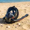 A full-face snorkel mask is lying on a sandy beach with the ocean in the background. Podgląd zdjęcia produktu