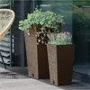 Two tall planters with plants on a wooden deck next to a chair and a glass door. Produktbild-Vorschau